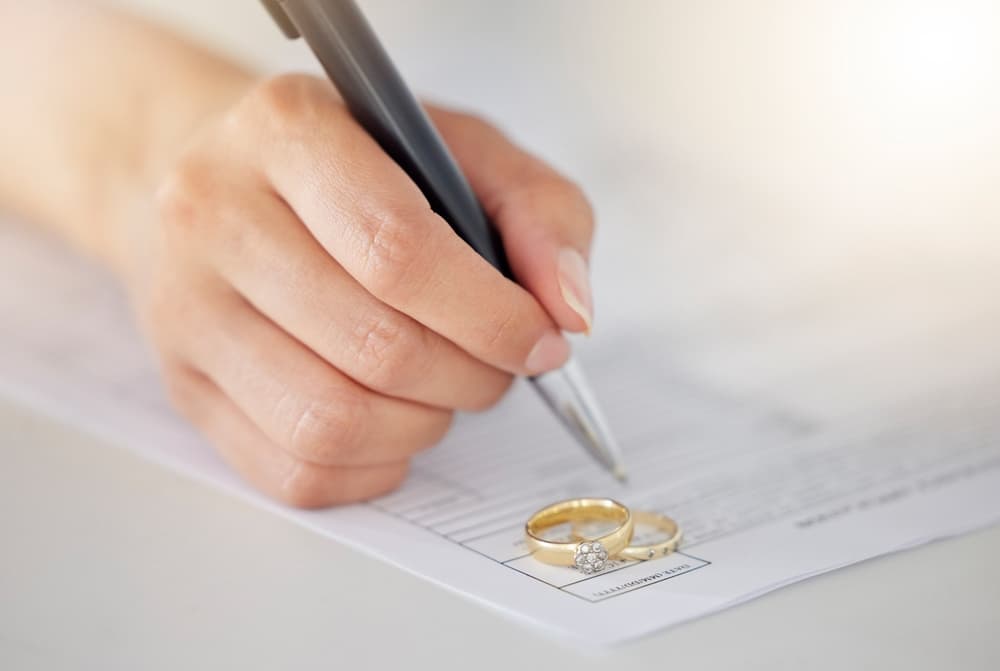 Woman hand signing legal divorce documents, deal or paper contract in a lawyer office with ring placed on table.