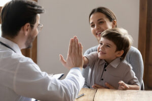 A smiling young boy gives a high-five to a male professional, possibly a doctor or attorney, while sitting beside his mother in a warmly lit office setting, suggesting a positive outcome or agreement.