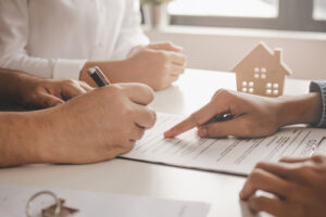 Close-up of hands signing a legal document on a desk, with a small wooden house model in the background, symbolizing a real estate or legal agreement.