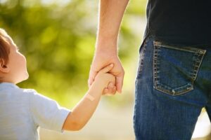 A young child with a small bandage on their arm holds hands with an adult, likely a parent, as they walk outdoors on a sunny day, symbolizing care, safety, and connection.