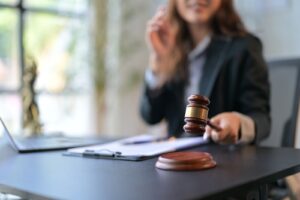 Female lawyer in a black blazer holding a gavel at her desk with legal documents and a laptop, symbolising courtroom authority and legal decision-making.