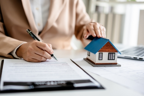 A person signs real estate documents at a desk while holding a small model house, symbolizing property and legal agreements.