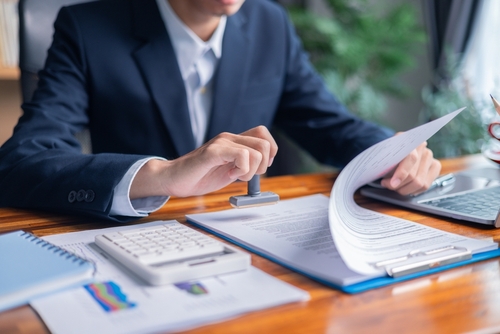 Business professional in a suit stamping official documents at a desk with a calculator, paperwork, and charts, representing legal or financial approval.